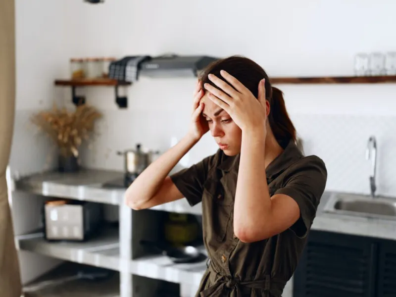 Mujer joven en la cocina con gesto de preocupación, llevándose las manos a la cabeza, sugiriendo dolor de cabeza o malestar.