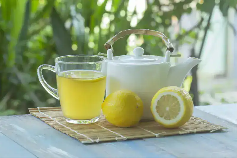 Taza de cristal con agua caliente y limón junto a una tetera blanca y limones frescos sobre un salvamanteles de bambú, con fondo de vegetación verde