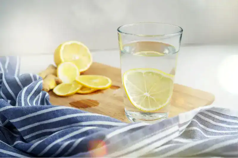 Vaso de agua con rodaja de limón junto a una tabla de madera con limones cortados y jengibre fresco, sobre un mantel de rayas azules y blancas