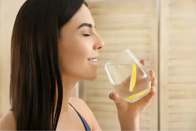 Mujer joven de cabello oscuro bebiendo un vaso de agua con limón, de perfil, con fondo de persiana de madera clara