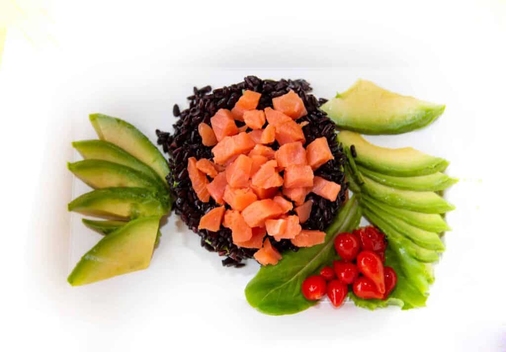 Plato de arroz negro Venere con dados de salmón ahumado, acompañado de láminas de aguacate, hojas de lechuga y tomates cherry, visto desde arriba
