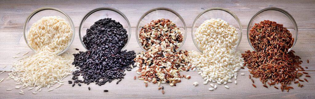 Cinco variedades de arroz en cuencos de cristal volcados sobre superficie de madera: arroz blanco de grano largo, arroz negro, arroz salvaje mixto, arroz redondo blanco y arroz rojo