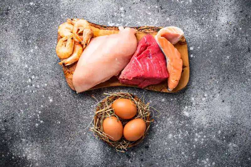 Carne de res, pollo, salmón y camarones sobre una tabla de madera, junto a huevos frescos en un nido de paja