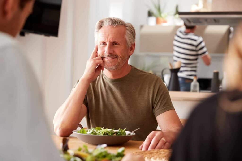 Hombre sonriente sentado a la mesa con un bol de ensalada, una comida consciente que cuida el aporte de proteínas.